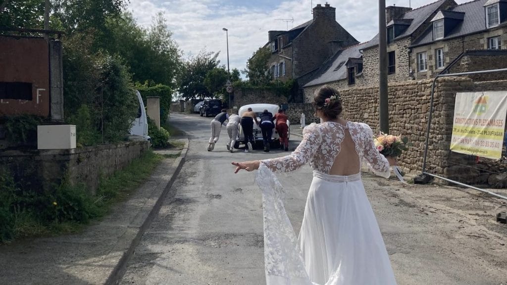 La mariée regarde les invités pousser la voiture ancienne dans une rue de Beaussais-sur-Mer